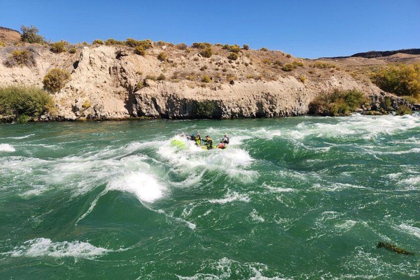 A river with white water rapids, a group of people rafting, and a rocky hillside in the background. 2