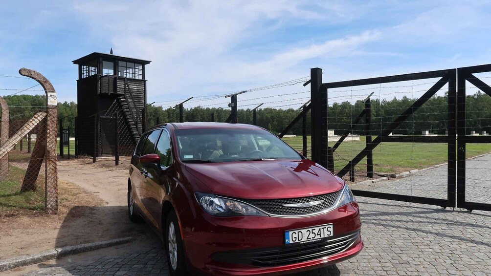 Punainen auto oli pysäköitynä portin lähelle. Portissa oli piikkilankaa, ja taustalla näkyi vartiotorni sekä hautausmaa. 2