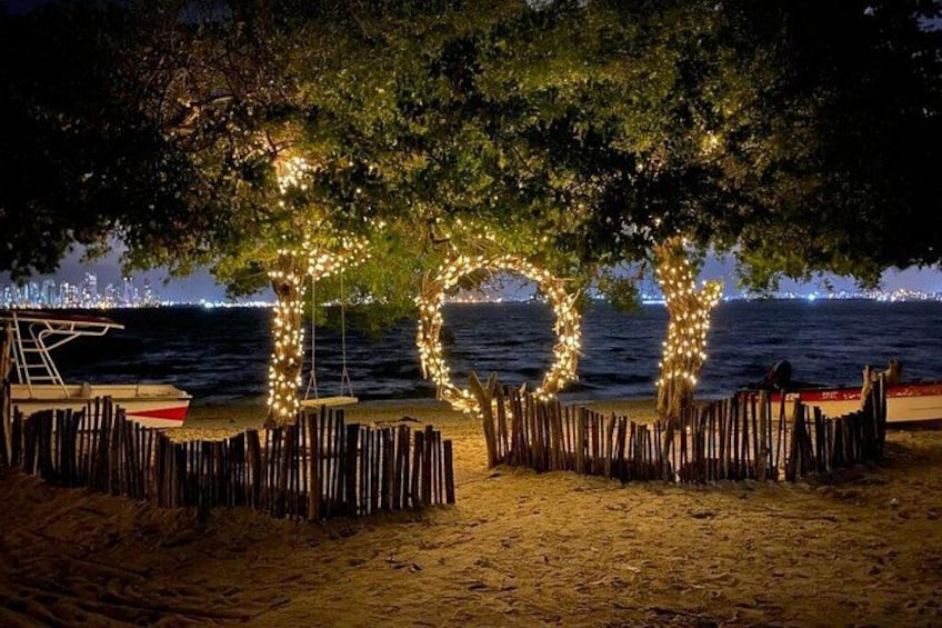 A beach at night with illuminated letters spelling 'LOVE' under trees, a boat, and a wooden fence. 2