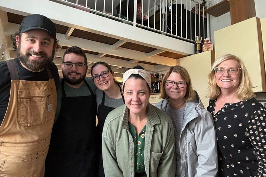 Un grupo de personas de pie en un interior, algunas con delantales, en una habitación con una escalera y armarios de cocina al fondo. 3