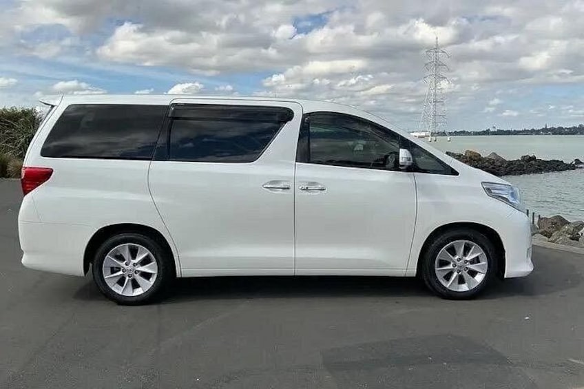 A white minivan parked on a paved surface with a body of water and a power line tower in the background. 4