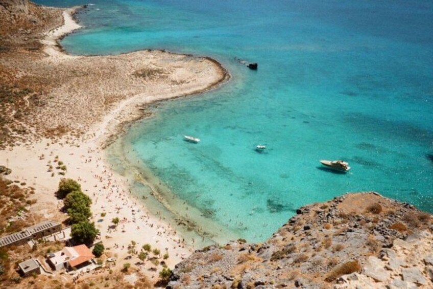 A coastal landscape with clear turquoise waters, a sandy beach, rocky terrain, and a few boats. 2