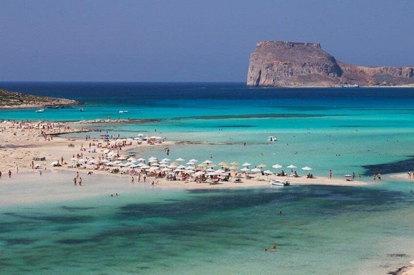 A beach with clear turquoise waters, a rocky outcrop in the distance, and numerous umbrellas and people. 4