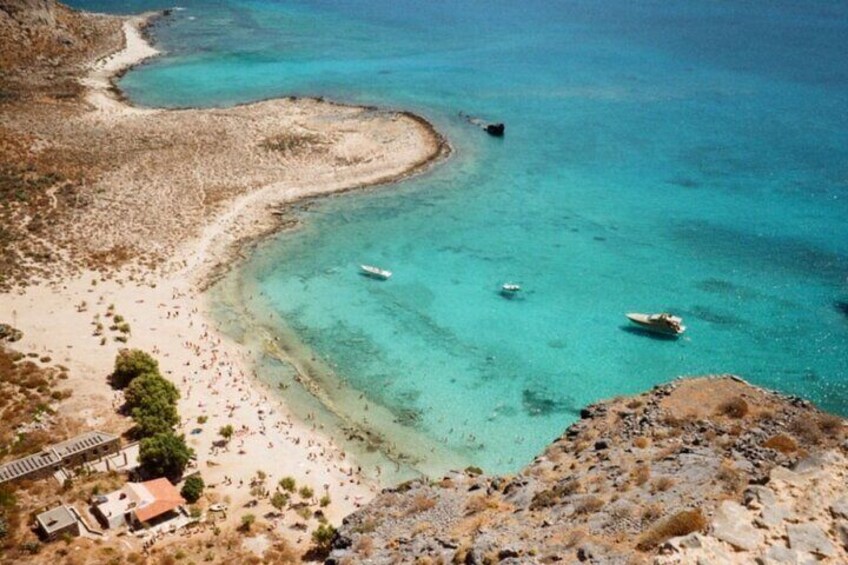 A coastal landscape with clear turquoise waters, a sandy beach, rocky terrain, and a few boats. 3