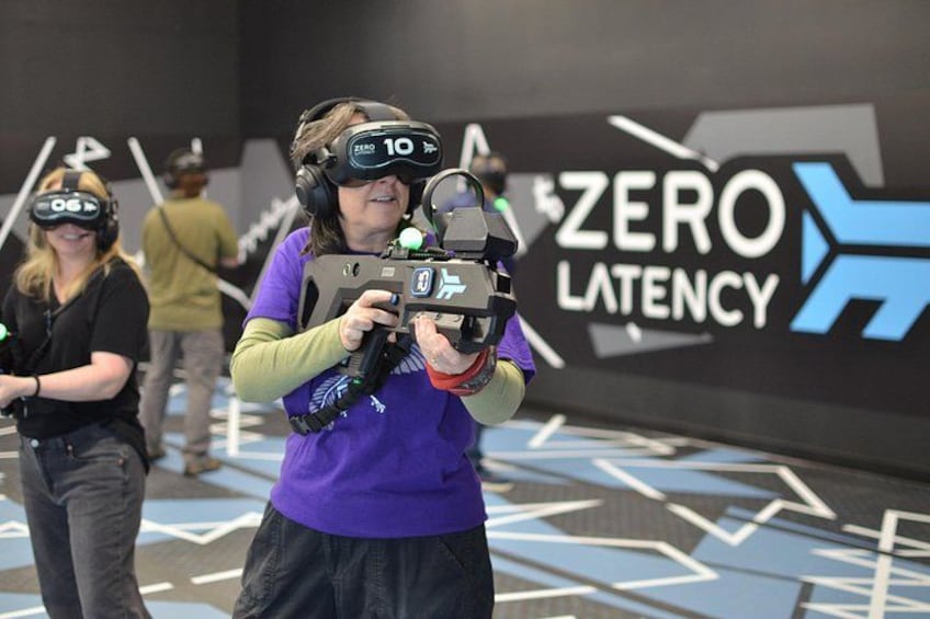 Una persona usando un casco de realidad virtual y sosteniendo un control de realidad virtual en una habitación con un letrero que dice 'ZERO LATENCY'. 5