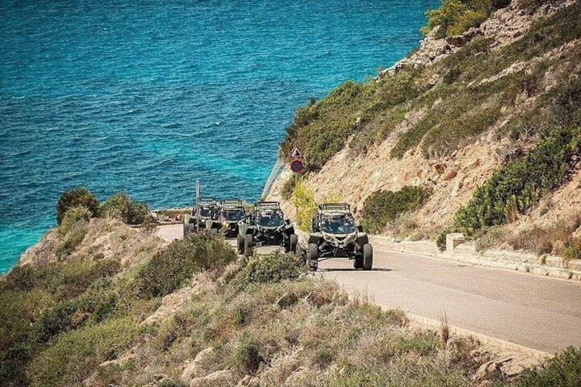 Un corteo di veicoli fuoristrada su una strada costiera con un precipizio ripido su un lato e un mare blu cristallino sullo sfondo. 1