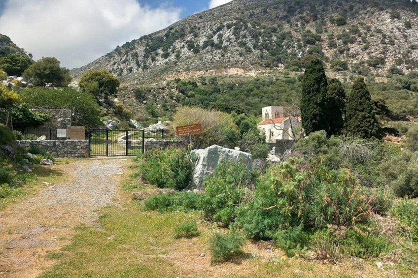 Paesaggio rurale con un sentiero sterrato, un muro in pietra e una chiesa circondata da vegetazione e colline. 5