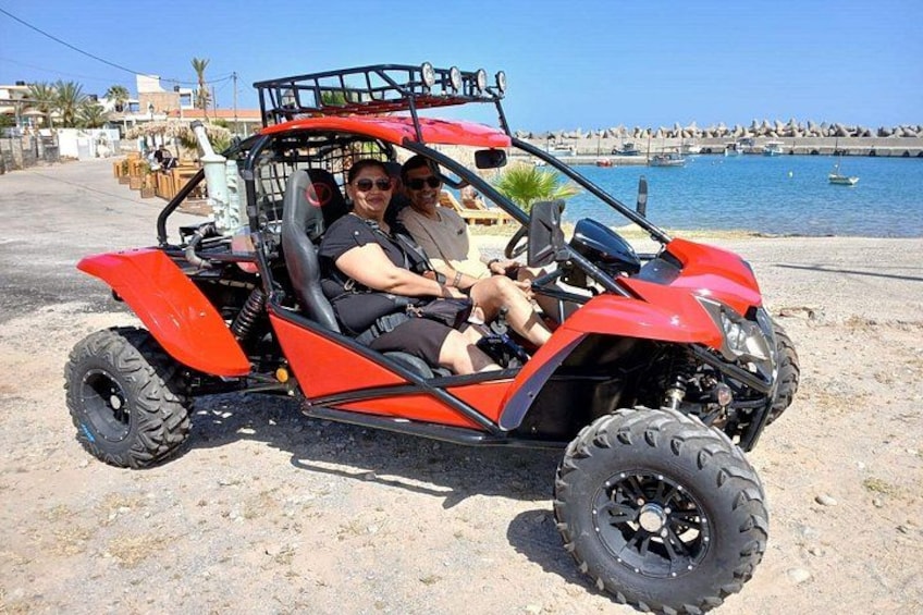 A red off-road vehicle parked on a sandy beach with a body of water and a rocky shoreline in the background. 5