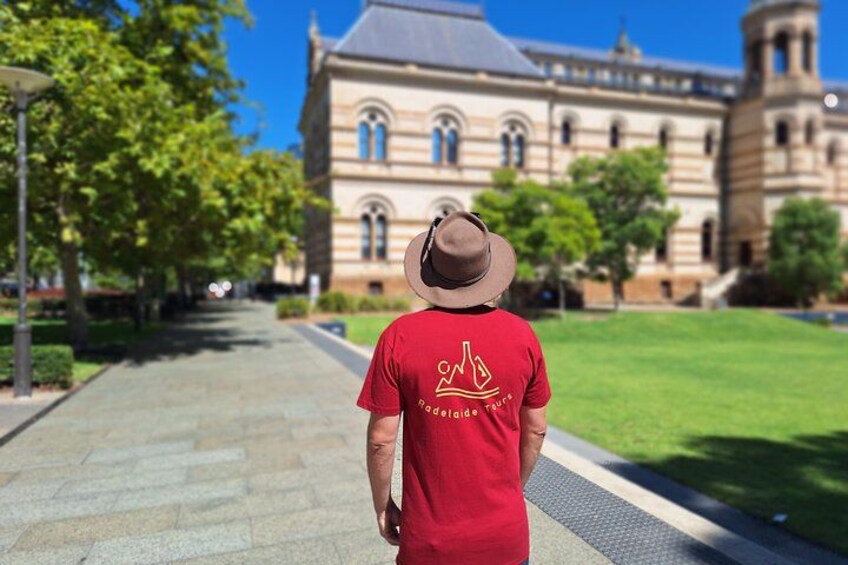 A person in a red shirt with a logo, walking on a paved path towards a large building with arched windows and a clock tower. 2