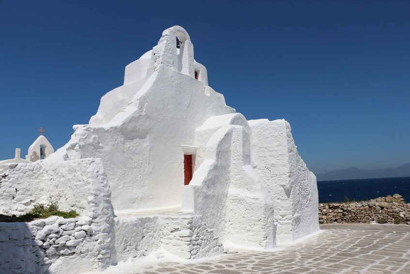 A white-washed building with a red door and a cross on top, set against a clear blue sky. 5