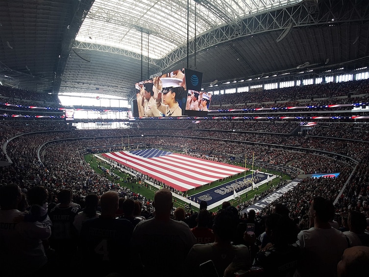 A large stadium filled with spectators, a large American flag on the field, and a scoreboard displaying images. 1