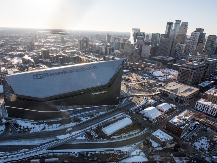 Ein großes Stadion, auf dessen Dach 'US Bank Stadium' steht, umgeben von einer Skyline mit hohen Gebäuden und Schnee auf dem Boden. 2