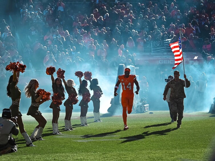En fotbollsspelare i orangefärgad tröja med nummer 14 springer på planen, omgiven av hejaklack och en militär som håller en amerikansk flagga. 4