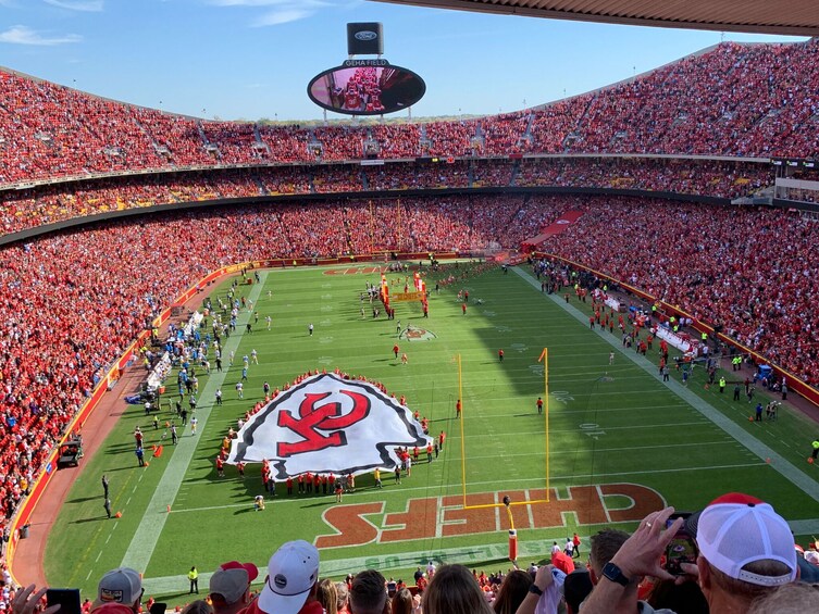 A football stadium filled with spectators, a large logo on the field, and a scoreboard. 1