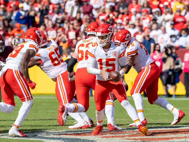 A football player in a white and red uniform holding a football, surrounded by teammates in similar uniforms. 5