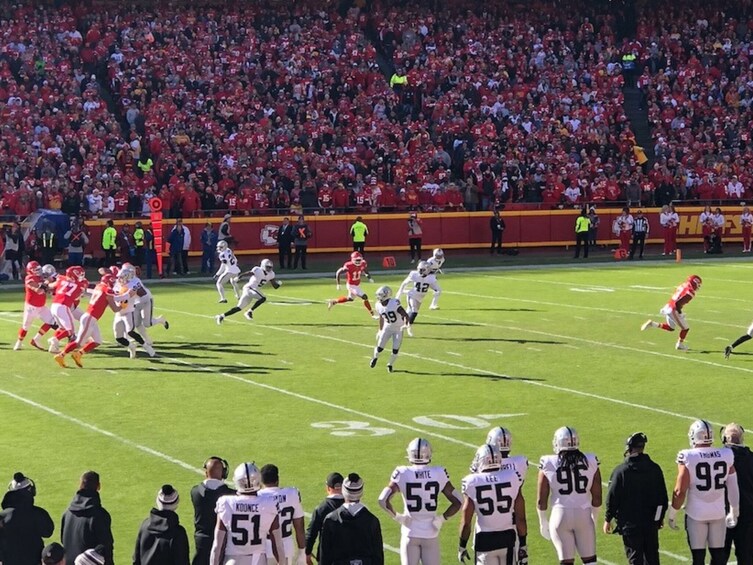 A football game with players in red and white uniforms on the field, a crowd in the background, and a sideline with coaches and staff. 3