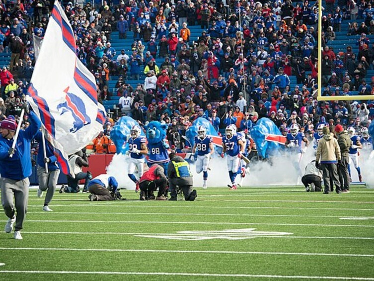 A football team on the field with players in blue and white uniforms, a crowd in the background, and a flag being carried. 1