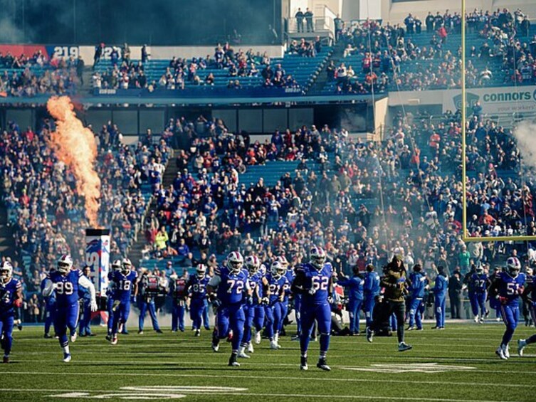 A football team in blue uniforms with numbers visible, running on a field with spectators in the background. 3