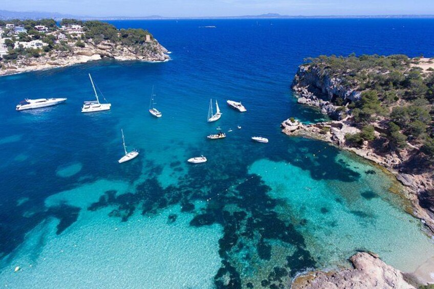 A coastal area with clear turquoise waters, several sailboats anchored near the shore, and rocky terrain with sparse vegetation. 3