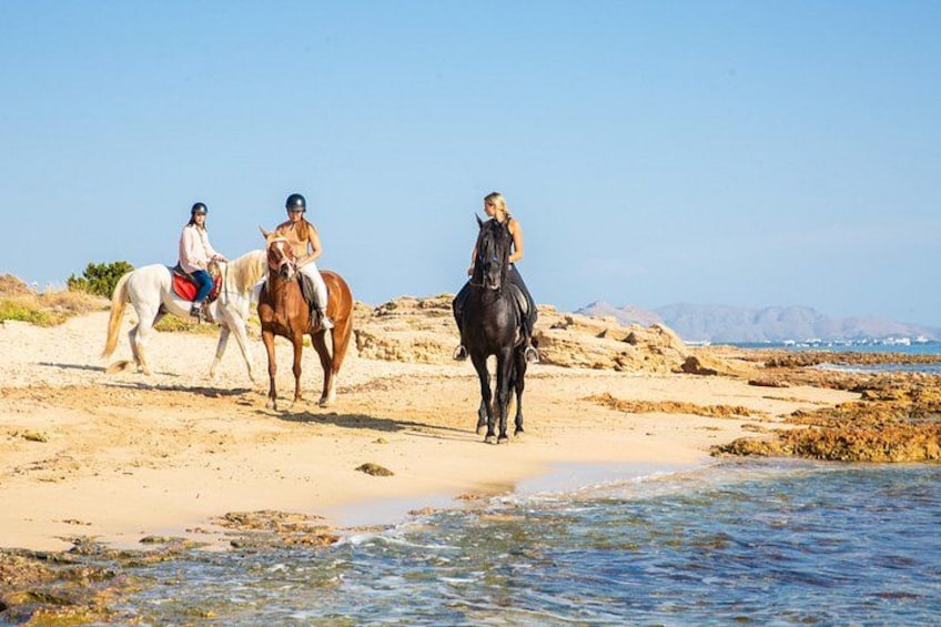 Tre personer rider på heste på en strand med en stenet kystlinje og en klar himmel. 2