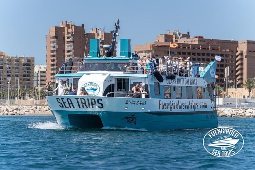 Una barca bianca e blu chiamata 'SEA TRIPS' con la scritta 'GLASS BOTTOM BOAT' sul fianco, che naviga sull'acqua con edifici sullo sfondo. 4