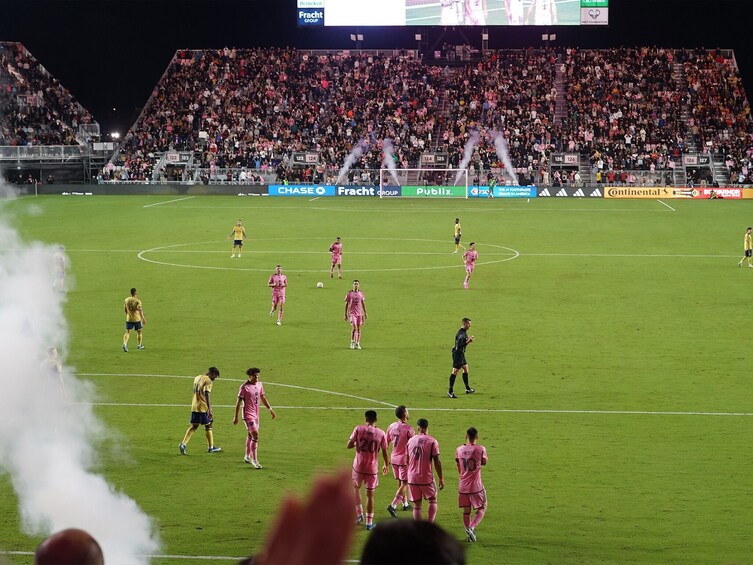 A soccer stadium filled with spectators, a soccer field, and players in pink and yellow uniforms. 4