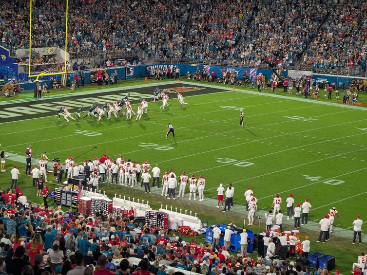 A football stadium with players and staff on the field, spectators in the stands, and a large crowd. 4