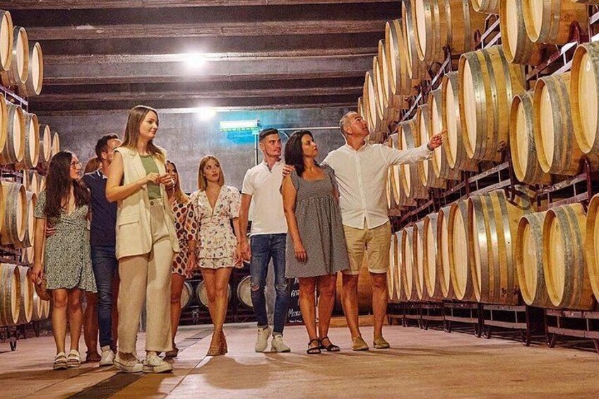 A group of people in a wine cellar with rows of wine barrels. 1