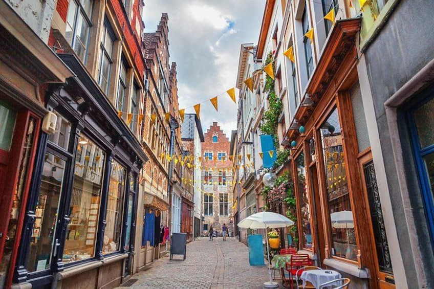 Una calle adoquinada flanqueada por edificios históricos, algunos con amplias ventanas y otros con contraventanas de madera. Banderines amarillos en forma de triángulo cruzan la calle, y se observan algunas mesas y sillas al aire libre. 2
