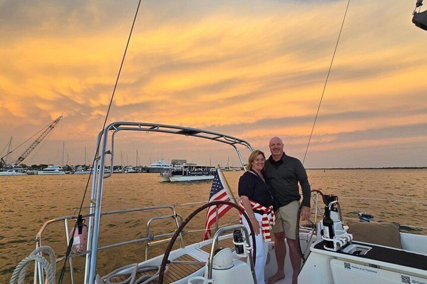 A couple on a sailboat with an American flag, a sunset, and other boats in the background. 2