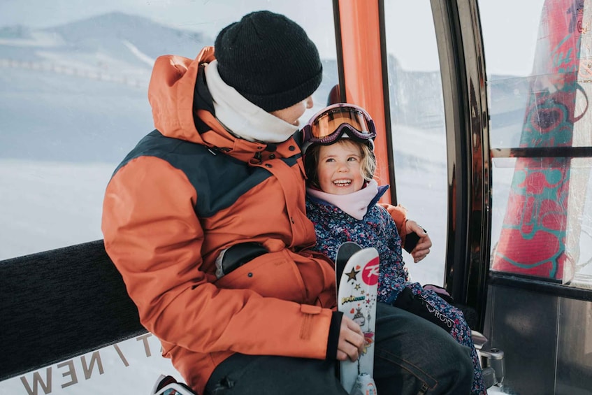 A person in an orange jacket and black beanie sitting next to a child in a blue jacket and helmet, holding a snowboard. 3