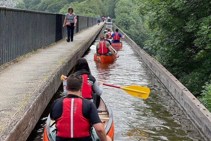People in canoes on a narrow waterway, a bridge in the background, and greenery along the sides. 1