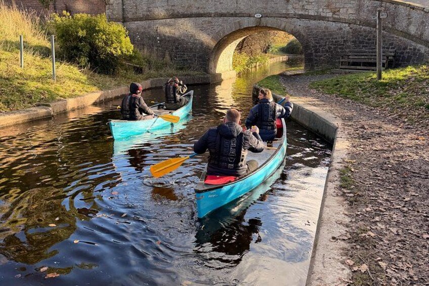 People in canoes on a narrow waterway, a stone bridge in the background, and a grassy bank on the side. 4