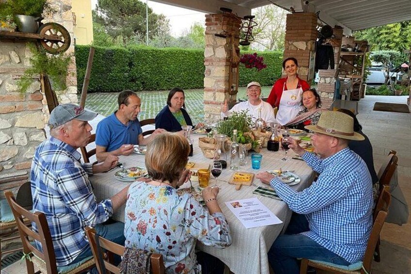 Eine Gruppe von Personen speist draußen an einem Tisch, im Hintergrund eine Backsteinarchitektur und Grünpflanzen. 1