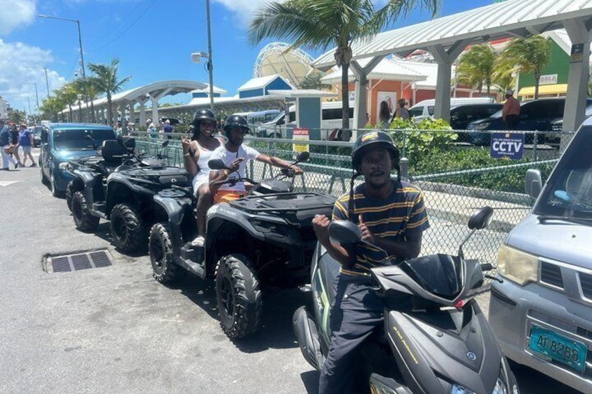 A group of people on ATVs and a scooter parked in a lot with a CCTV sign and palm trees in the background. 3