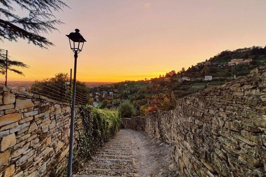 A stone pathway leading uphill, flanked by stone walls, with a street lamp and a sunset in the background. 2