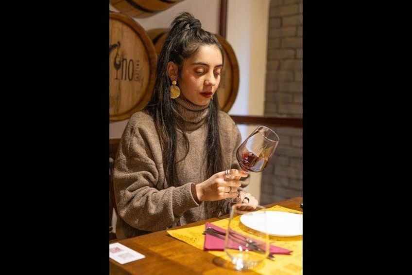 Una persona seduta a un tavolo con un bicchiere di vino, circondata da botti di legno e un muro di mattoni. 5