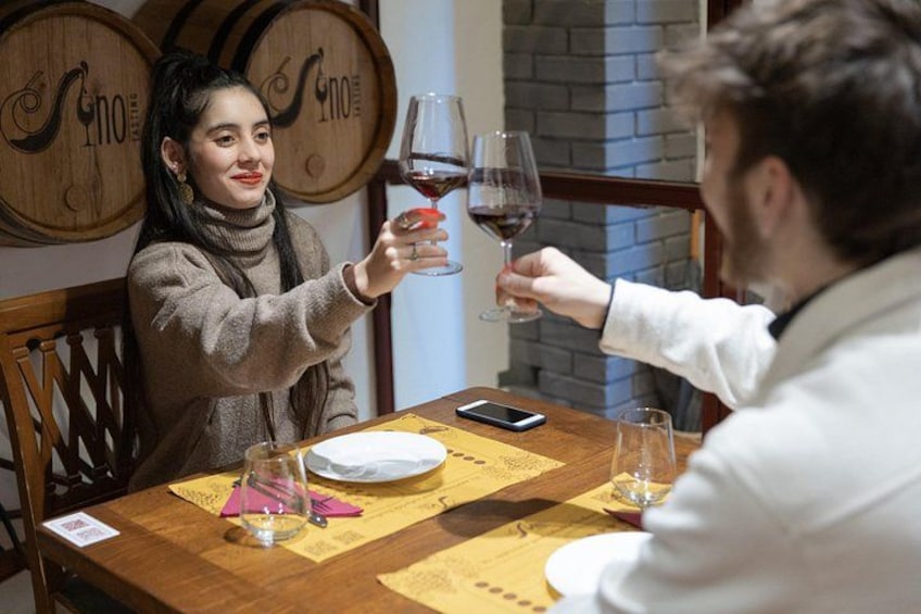 Due persone brindano con bicchieri di vino a un tavolo di legno, con un muro di mattoni sullo sfondo. 2