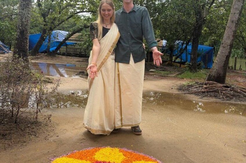 A person in a beige saree standing on a dirt path with a colorful rangoli in the foreground. 5