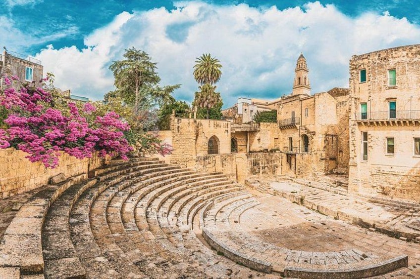 Un antico anfiteatro con gradinate in pietra e un palco circolare, circondato da edifici storici e alberi in fiore. 3