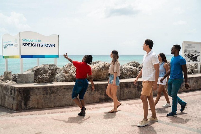 A group of people walking along a waterfront promenade with a sign reading 'Welcome to Speightstown.' 2