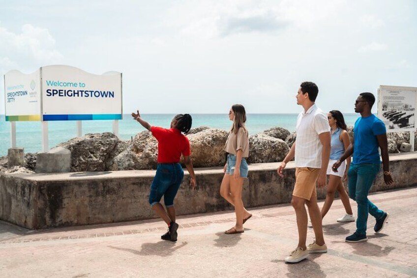 En gruppe mennesker går langs en strandpromenade med et skilt, der siger 'Welcome to Speightstown'. 2