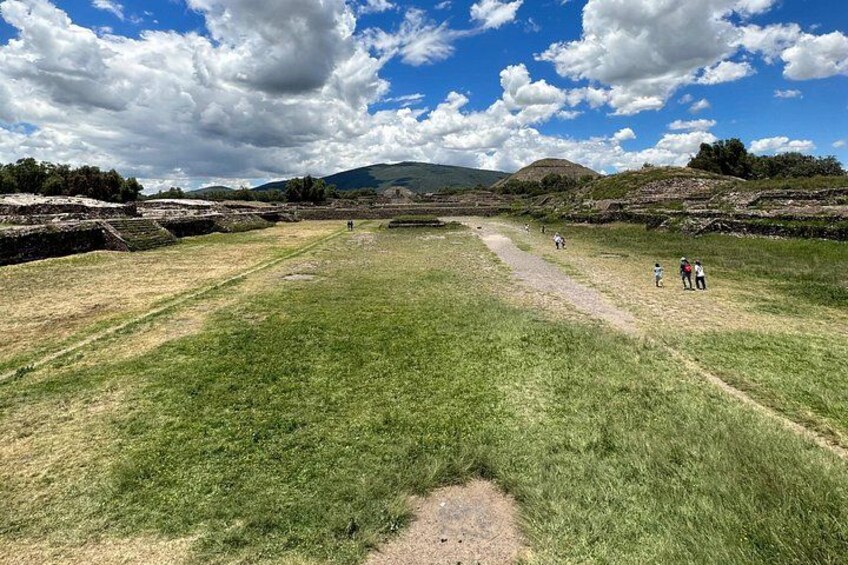 A grassy field with a dirt path, a few people walking, and a hilly landscape in the background. 2