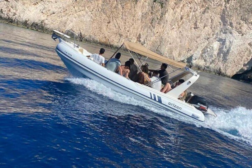 A white motorboat with a canopy, named 'erizon,' moving through blue water with a rocky coastline in the background. 2