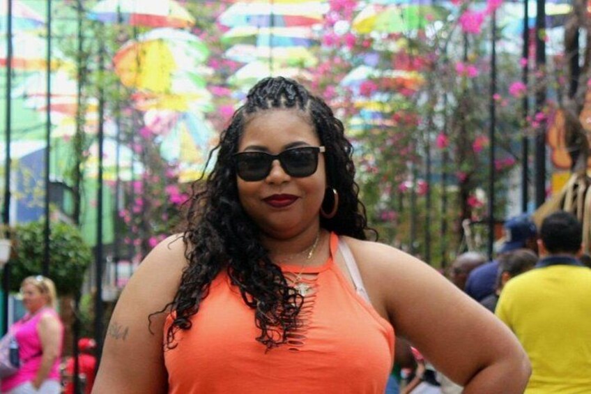 Iemand met een oranje top en een tatoeage op hun arm, staand in een open landschap met kleurrijke parasols en groen op de achtergrond. 4