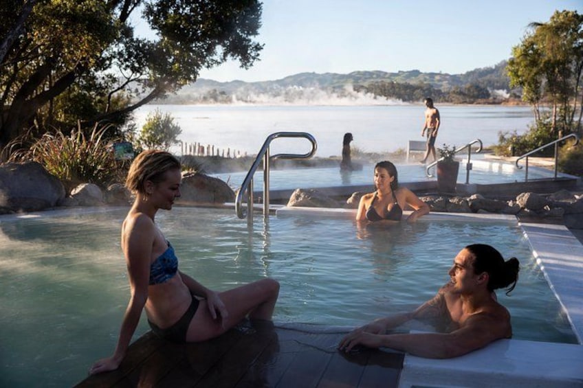 A hot spring with people relaxing, a wooden deck, and a lake with mountains in the background. 2