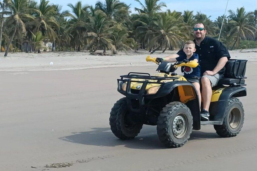 A man and a child on a yellow ATV on a sandy beach with palm trees in the background. 1