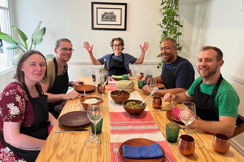 A group of people sitting around a wooden table with various bowls, glasses, and utensils. 1