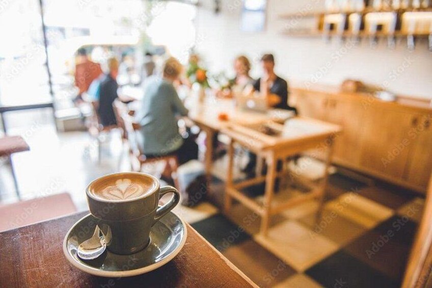 A coffee cup with latte art on a saucer, a wooden table, and a checkered floor in a cafe with people in the background. 2