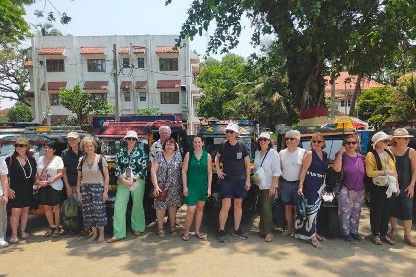 A group of people standing outdoors in a sunny area with a building and trees in the background. 3